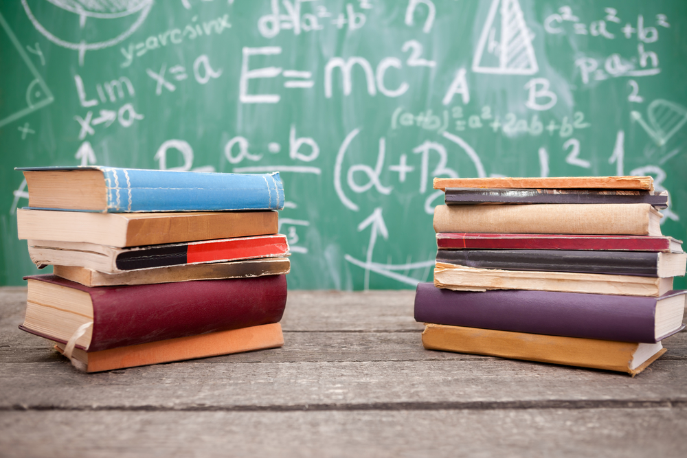 Two stacks of colorful school books on a wooden desk in front of a chalkboard covered in math equations